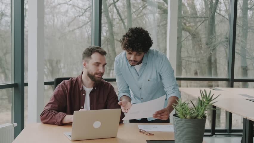Two diverse employees in casual planning working talking together at contemporary office using laptop. Multiethnic executive team discussing financial report sitting at table. | Shutterstock HD Video #1100786133