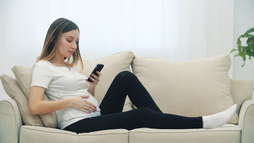 4k slowmotion video of pregnant woman sitting on white sofa with phone. | Shutterstock HD Video #1100788147