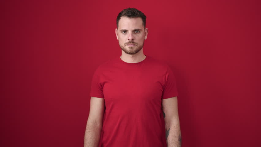 Young caucasian man standing with serious expression taking t shirt off over isolated red background
