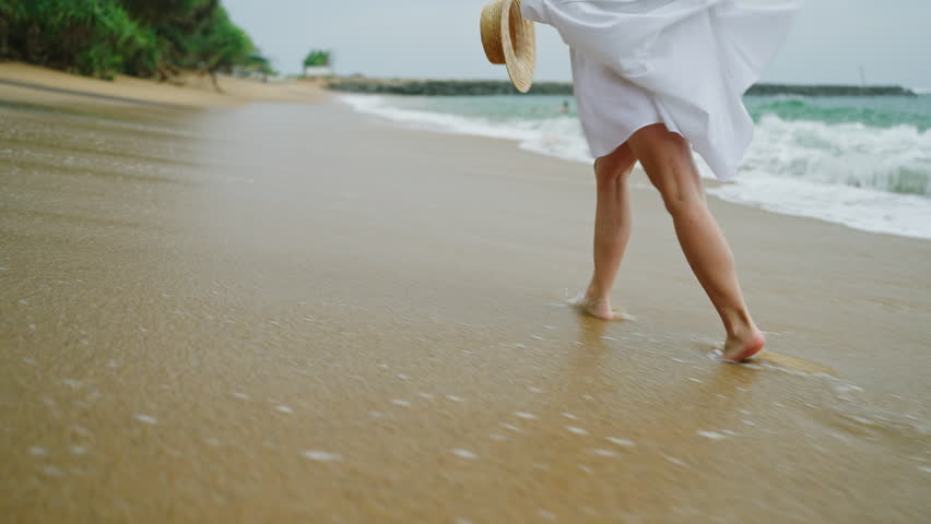 Slim female legs and feet walking along sea water waves on sandy beach. Pretty woman walks at seaside surf wearing white shirt carrying a straw hat gets splashed with waves. Girl running along shore
