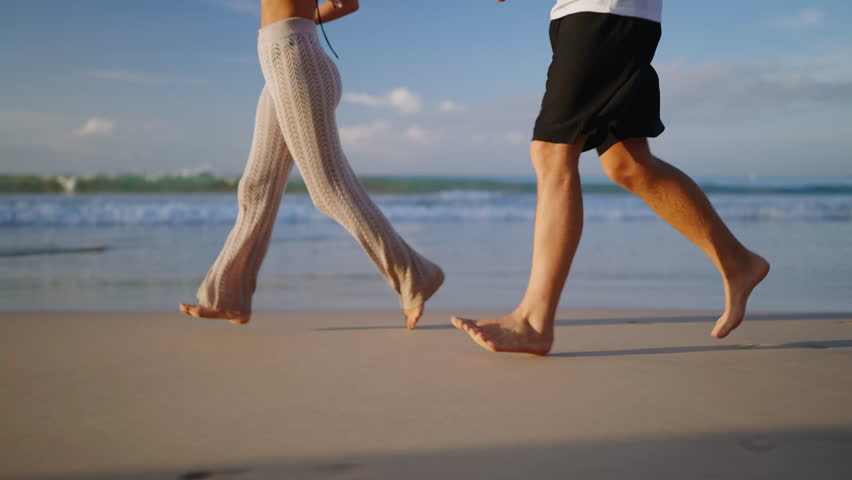 Legs of happy couple holding hands and walking on the beach together enjoying summer side view. Feet of heerful boyfriend and girlfriend relaxing and taking a walk at the seaside at sunrise side shot.