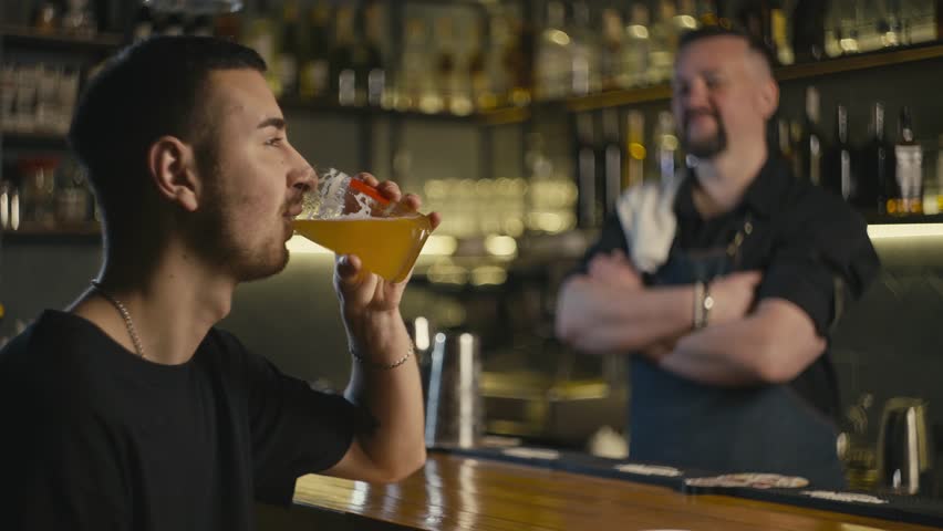 Satisfied man drinking beer at bar, bartender smiling in background. A great end to a working day. High quality 4k footage | Shutterstock HD Video #1100795501