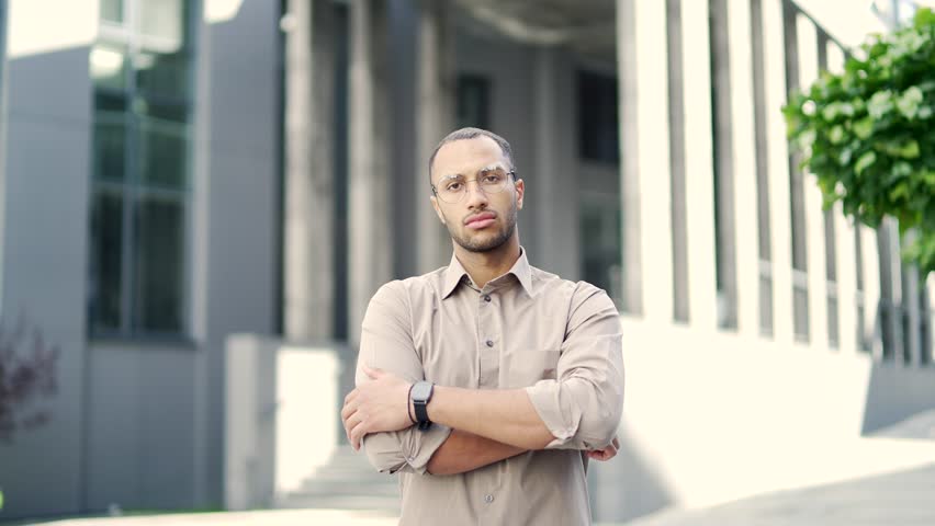 Portrait of a young adult man in a shirt and glasses seriously looking at the camera while standing on the street near an office building. Confident handsome mixed race male posing with crossed arms