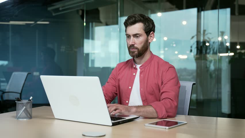 Handsome bearded employee businessman worker works on laptop in modern glass office. confident male business entrepreneur freelancer in red shirt typing browsing using computer on desktop indoor Royalty-Free Stock Footage #1100795895