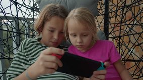 Close up shot of two siblings playing video games on a gaming console while seated together on a swing chair on the balcony. The older brother (12 years old) is helping and guiding his younger sister
