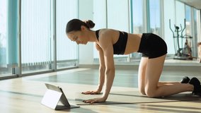 Beautiful sporty woman in sportswear doing plank with leg and hand raise while watching training video on tablet. Indoor studio shot illuminated by sunlight from window