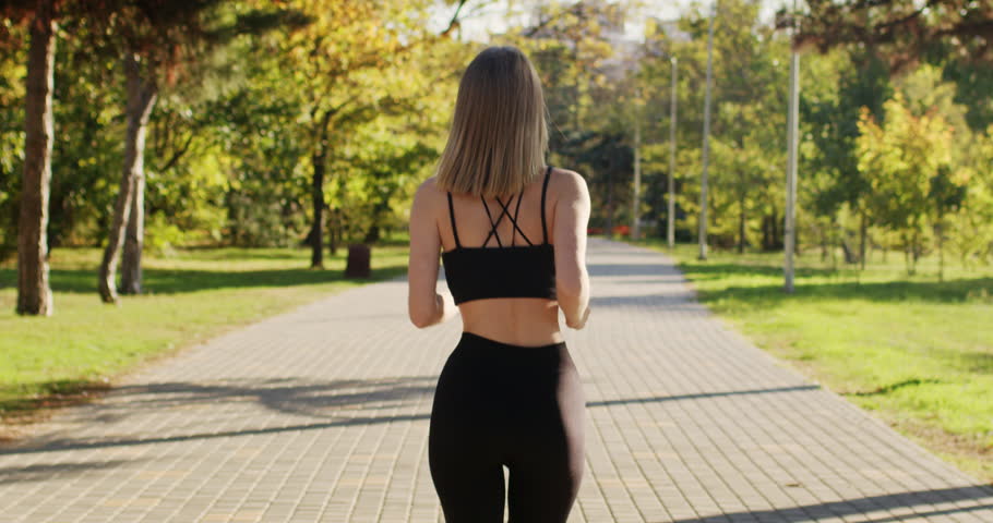 Blonde short-haired woman practices running in city park illuminated by sunlight. Young woman wearing black sports top enjoys jogging