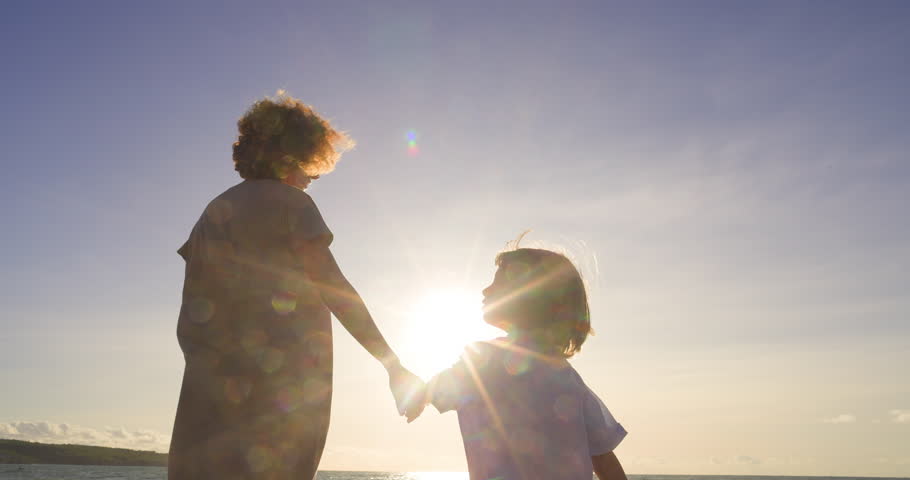 A son and mother walk in park holding hands. Glare from scorching sun in frame, where happy family of mother and child walk and smile, rejoicing. Bright warm summer days filled with love and joy.