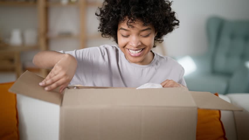 Happy satisfied african american woman customer receive carton box and open delivered parcel excited good purchase online shipping. Smiling happy consumer get post package look inside sit sofa home. Royalty-Free Stock Footage #1100824711