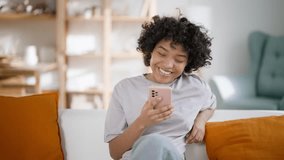 Smiling beautiful african american woman looking at telephone screen, reading text message with good news. Celebrating success online lottery win or shopping, internet communication. Home lifestyle.