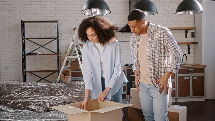 Happy young married couple, a wife and husband unpacking cardboard box with personal belongings, admiring family photo in a frame, standing together in their new own apartment. Moving into new house Royalty-Free Stock Footage #1100825829