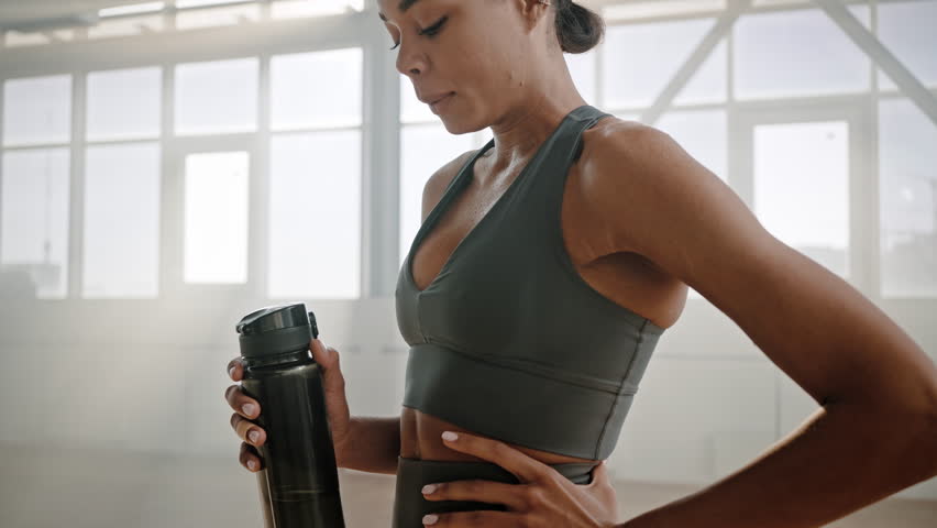 Multi-ethnic young athletic woman drinking water from sport bottle, relaxing and renewing the water balance after intense workout in smokey gym interior. Perspiring sportswoman resting after training Royalty-Free Stock Footage #1100825917
