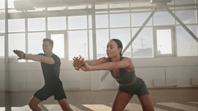 Slow motion video of young determined athletic team in tracksuit, doing side lunges and bending body with outstretched arms forward during fitness workout indoors. Stretching. Sport. Endurance concept