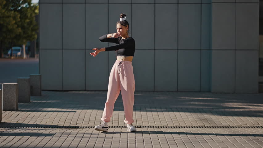 Young professional lady dancer wearing black blouse performs vogue moves against building. Stylish woman with tattoo on belly enjoys performance at sunrise | Shutterstock HD Video #1100826305