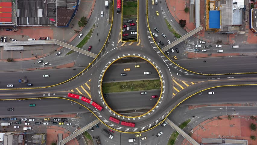 A bird's eye view shot with a 4K drone descending on a round point intersection with red articulated buses, taxis and other vehicles turning from Third street to 30th avenue in Bogotá, Colombia. Royalty-Free Stock Footage #1100837601