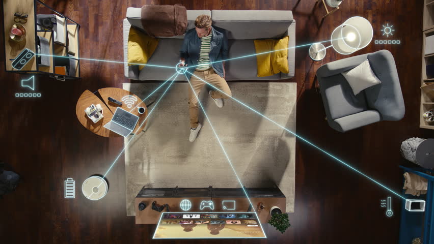 Top View Of Caucasian Man In the Loft Apartment Sitting Down on The Couch and Connecting Smartphone to Convenient Smart Home System. VFX Animation Visualizing Connected Devices. Laptop, TV, Speaker.