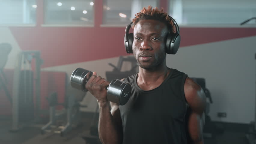 exercicing in gym with dumbbells, portrait of handsome black man lifting weights, training biceps, developing muscles of arms and shoulders, workout in fitness center, listen to music by headphones