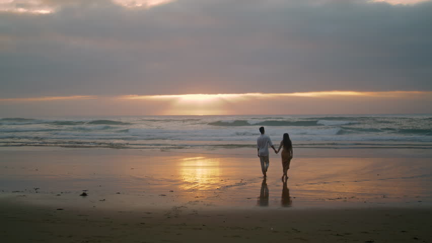 Affectionate newlyweds going sunrise sea shore. Loving woman holding man hand on morning sky beach. Married people watching ocean horizon back view. Love sweethearts enjoying romantic time together Royalty-Free Stock Footage #1100853907