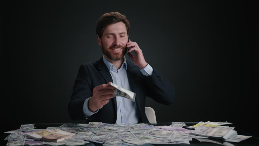 A man in a suit dropping money while talking with someone on the phone. He looks happy. The man is in black room. The m,oney is in a big pile. High quality 4k footage 