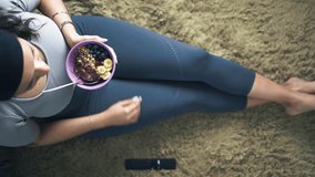 Latina woman relaxing after workout eating muesli and smoothie bowl while watching television in the floor of the living room. Top view video shot