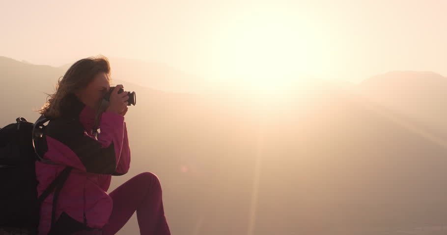A woman hiker photographer taking pictures sitting on the top of the mountain, against the sun background. Silhouette view