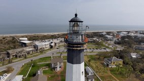 orbit aerial footage of the Tybee Island Light Station with vast ocean water, hotels and homes, lush green trees and grass and a sandy beach with cars driving on the street in Savannah Georgia USA