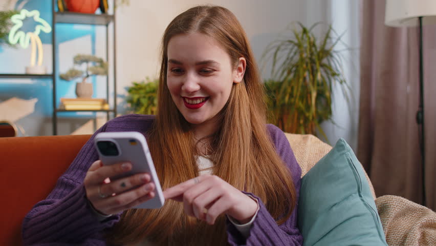 Woman sitting on sofa uses mobile phone smiles at home living room apartment. Portrait of young girl texting share messages content on smartphone social media applications online, watching relax movie | Shutterstock HD Video #1100866679