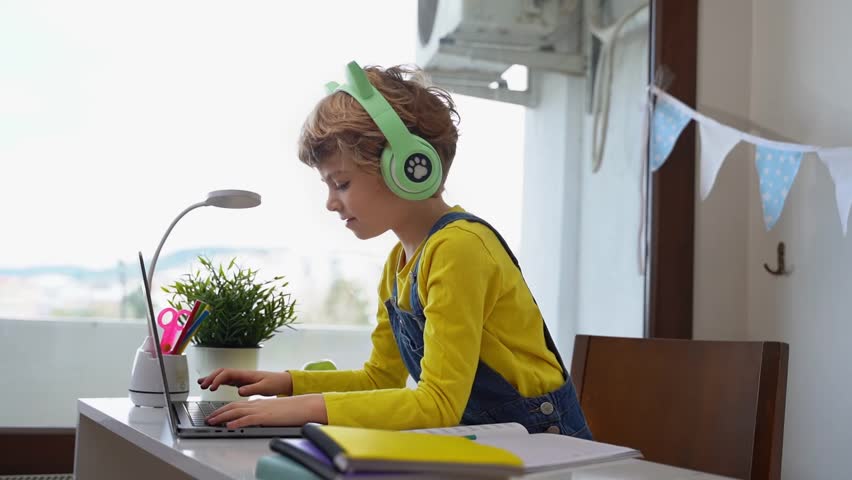 Cute smart caucasian primary School Child with headphones typing on the keyboard laptop. Online tutor teaching digital class, web lesson on computer at home. Virtual education. Schoolboy girl studying Royalty-Free Stock Footage #1100868595