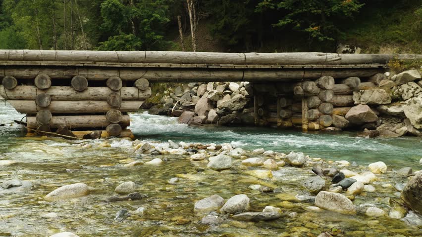 Powerful long wooden log bridge and clear beautiful mountain river flowing beneath it. The water in the river is turquoise in colour. Rocks lie on the bank. The beauty of nature