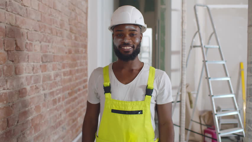 Construction worker in uniform put on safety goggles smiling at camera. Builder in overalls and hardhat put on glasses. Building, repair and renovation of house or apartment. Realtime Royalty-Free Stock Footage #1100874625