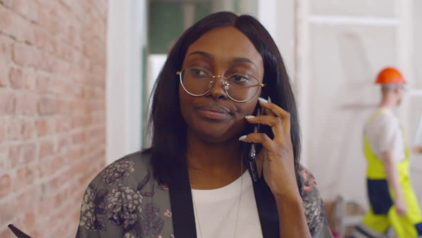African-American woman talk on smartphone at construction site. Female interior designer talk on phone with client about house renovation. Woman architect talk on cellphone. Realtime 