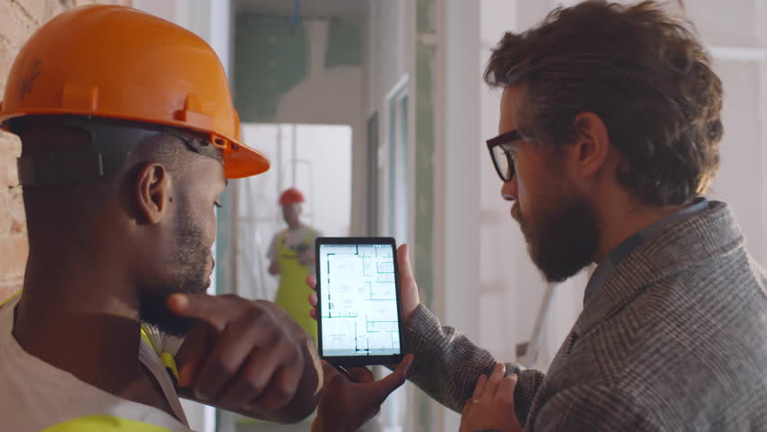 Home owner and builder discuss house blueprint on digital tablet. Back view engineer and architect study house plan on tablet pc. Man with construction worker use tablet for renovation. Realtime.  Royalty-Free Stock Footage #1100874633