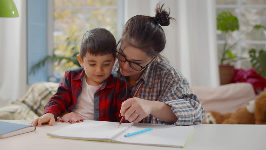 Cute kid learning writing with young mom tutor. Adult parent mother teaching school boy helps with homework. Mom and son studying sitting at home table. Children education concept. Realtime Royalty-Free Stock Footage #1100874651