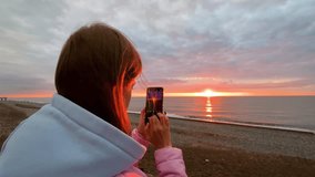 Woman is standing on the sea shore, holding smartphone and taking photo or shooting video of the Black Sea, warm sunset sky - slow motion. Freedom, photography and summer concept