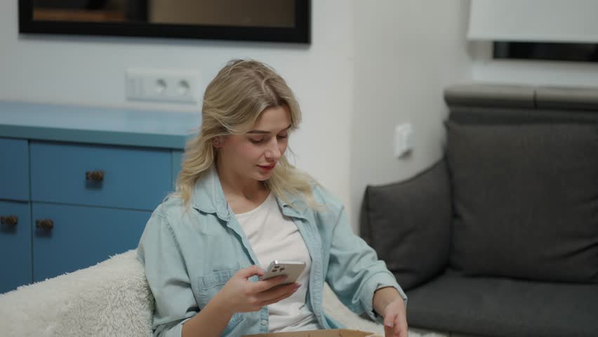 Bored girl sitting on sofa at modern living room and eating tasty pizza while use mobile | Shutterstock HD Video #1100878831