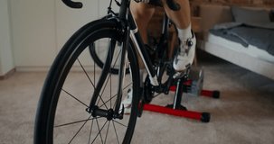 Young sporty man cycling in modern home interior using bicycle holding station, dynamic indoor video, frame movement from male legs to his face