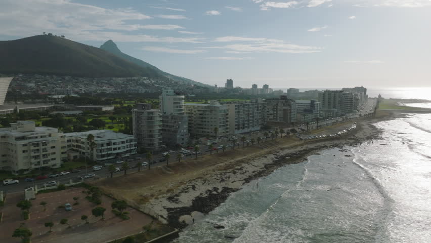 Row of multistorey buildings along sea coast. Waves washing shore. Water surface reflecting bright sunshine. Cape Town, South Africa