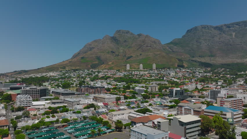 Aerial view of various buildings in residential borough. High and rocky mountain ridge against clear blue sky in background. Cape Town, South Africa