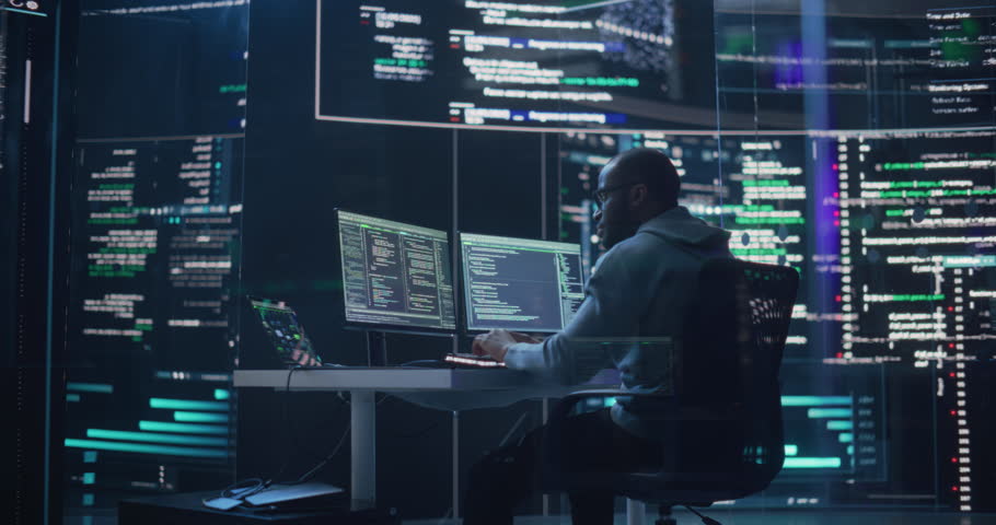 Medium Shot of a Man Working as a Developer, Surrounded by Big Screens Displaying Lines of Code in a Monitoring Room. Black Male Programmer Using Desktop Computer, Analysing Data, Creating AI Software Royalty-Free Stock Footage #1100883655