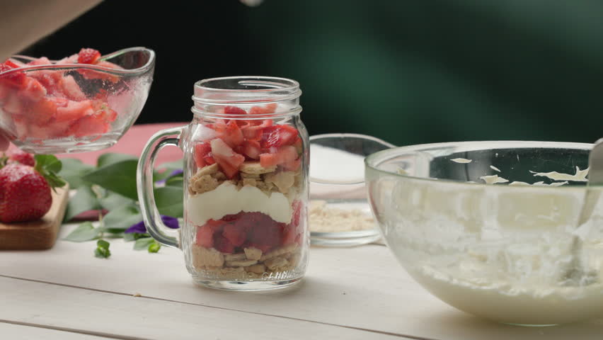 Assembling dessert - adding macerated chopped strawberries to jar with layers of whipped cream and crushed crackers; homemade dessert | Shutterstock HD Video #1100886657