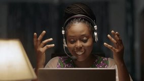 Young African Lady Wearing Headset behind laptop computer, video conferencing, female freelancer working online from home.