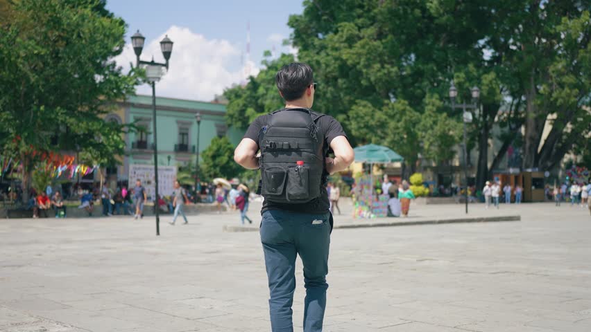 Slow motion male traveler walking outside old cathedral, zoom in pan around view of Hispanic men with sunglasses and backpack on sunny day in Oaxaca, Mexico