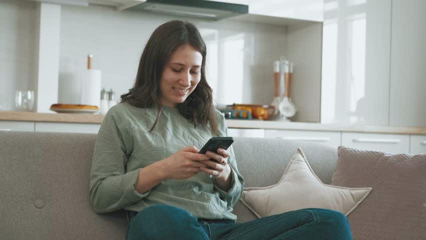 Footage of young cheerful woman sitting on sofa and typing in mobile phone at home | Shutterstock HD Video #1100892023