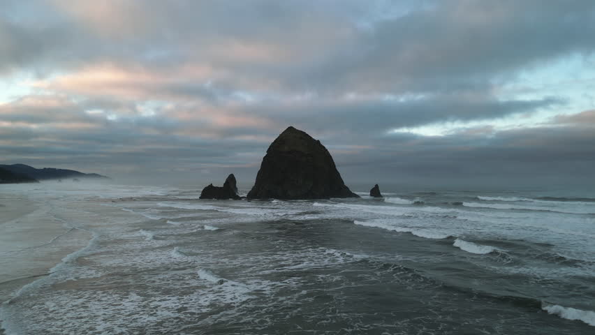 Haystack Rock in Cannon Beach, Oregon, United States. Dark rock formation in water. Cloudy dramatic weather. Waves splashing surf on sandy beach. High quality UHD 4k footage