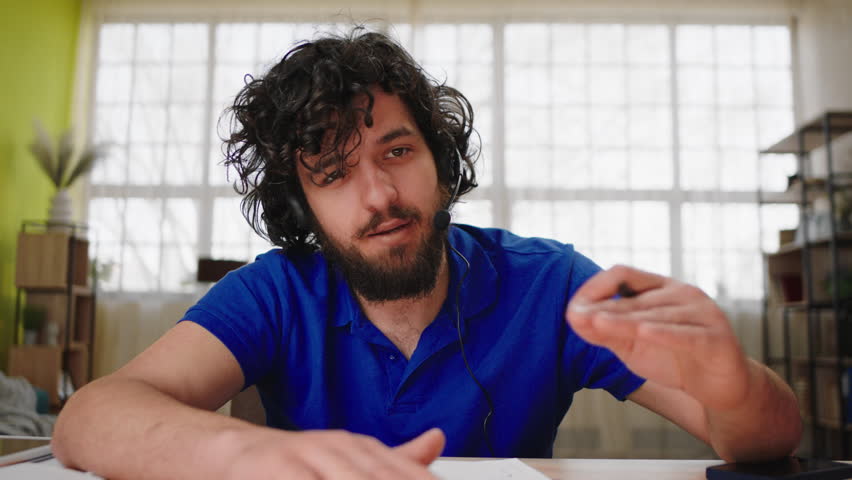 In front of the camera charismatic young man support centre worker discussing with client online from the headset he explains something