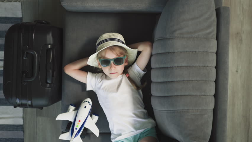 Small girl in straw hat with hands behind head relaxes at home before departure. Upset preschooler in sunglasses prepared for vacation upper closeup Royalty-Free Stock Footage #1100902011