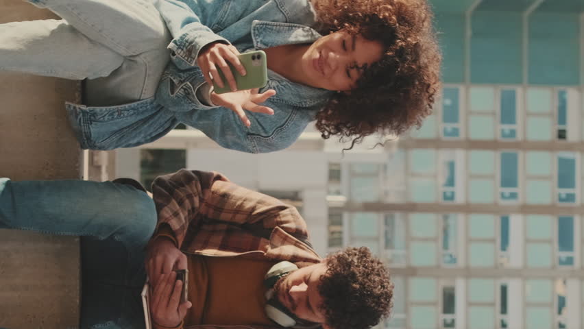 VERTICAL VIDEO, Couple of students are using mobile phones and talking while sitting outside. | Shutterstock HD Video #1100905049