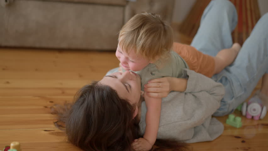Happy carefree mother and cute small child son tickling laughing having fun lying on floor and playing, smiling mum enjoying bonding cuddling hugging spending time with little funny kid at home