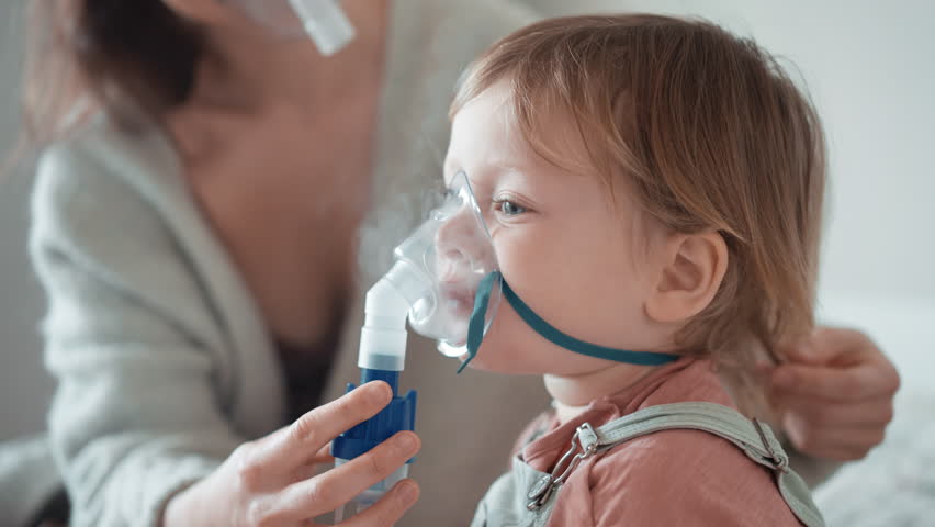 Asian mother helping sick daughter use nebulizer while embracing her on couch at home. Woman makes inhalation with equipment to toddler boy. Ill child lying on couch having respiratory illness helped | Shutterstock HD Video #1100907715