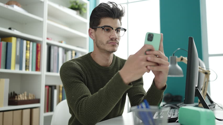 Young hispanic man student using smartphone sitting on table at library university